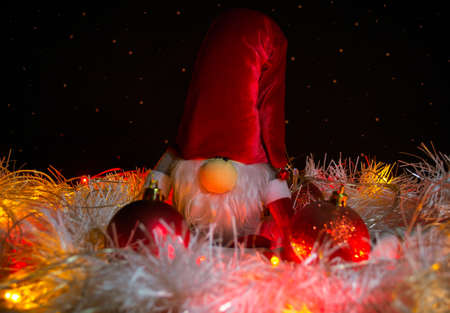 Toy New Year's gnome surrounded by red Christmas tree decorations, white tinsel and light garlands on the background of imitation of the starry skyの写真素材