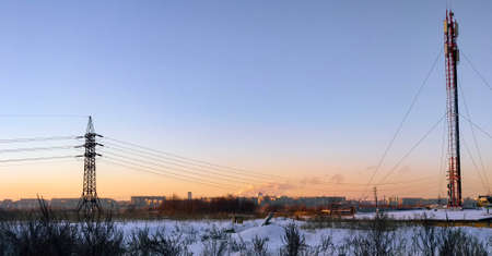 A power line pole and a radio communication tower stand in a winter field among the snow against the background of the evening sky and the city under the rays of the setting sunの写真素材