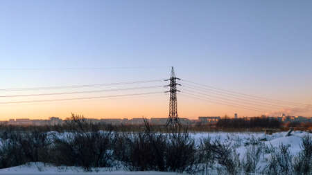 A power line pole stands in a winter field against the background of the evening sky and the city under the rays of the setting sunの写真素材