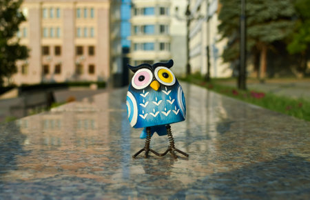 On polished granite, a surprised blue owl with multi-colored eyes stands close-up against the background of buildings in bokeh. Toy illuminated by evening sunlightの写真素材