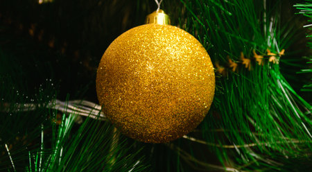 One toy in the form of a golden ball hangs against the background of green branches of an artificial...