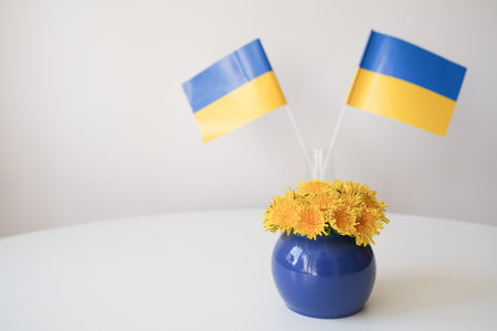 Yellow dandelions stand on a table, in a blue, round vase against the background of two flags of Ukraine. Space for your text. High quality photoの写真素材