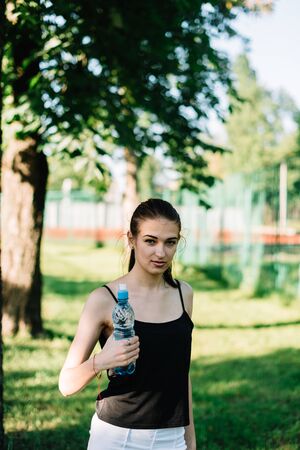 Girl athlete with a bottle of water in her hands. Summer sunny day in the park.の写真素材