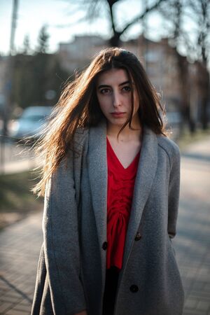 Girl in a red blouse and a gray cardigan on a blurry city background in early spring.の写真素材