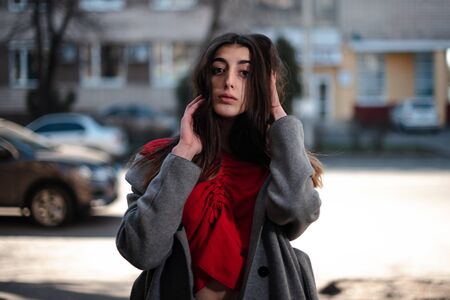 Girl in a red blouse and a gray cardigan on a blurry city background in early spring.の写真素材