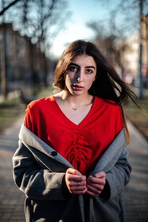 Girl in a red blouse and a gray cardigan on a blurry city background in early spring.の写真素材