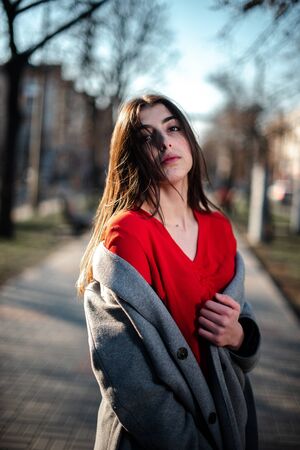 Girl in a red blouse and a gray cardigan on a blurry city background in early spring.の写真素材