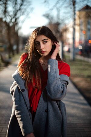 Girl in a red blouse and a gray cardigan on a blurry city background in early spring.の写真素材