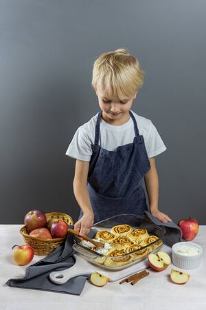 The process of glazing fresh baked cinnabon. Child smear buns with cream. On a white wooden table nearby are yaloks, which are also in the filling.の写真素材