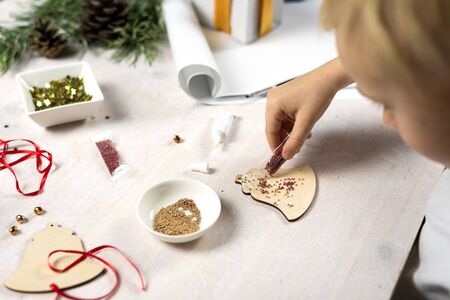 Cute boy thought about the craft. The child makes a Christmas tree decoration, holds a packaging with beads. He is sitting at a white table.の写真素材