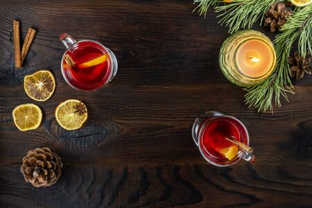 Two glass cups with mulled wine, with slices of orange and a stick of cinnamon, and burning candle in a jar are on a wooden brown table. Flat lay on a dark background with copy space in the center.の写真素材