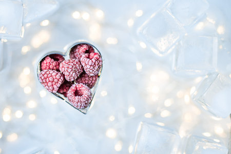 Frozen raspberries in the shape of a heart on a white-blue background with ice cubes and yellow...