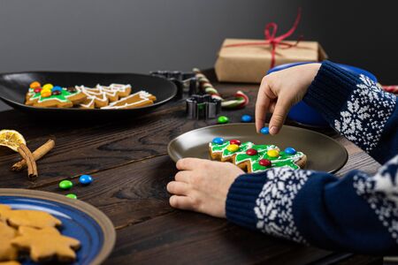 The child decorates with candys a gingerbread in the shape of a Christmas tree. Close up children's hands, lots of sweets on a wooden table.の写真素材