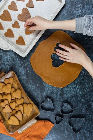 Womans hands take a gingerbread cookies, shape of hearts. Preparation for valentines Day. Beside is a baking tray and a wooden board with fresh pastries. Vertical flat lay cooking on dark blue table.の写真素材