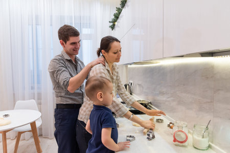 Mother and son are preparing cookies in the kitchen. Dad does a neck and shoulder massage to his wife while she cooks. Fun family tradition to cook together. Caring for loved ones.の写真素材