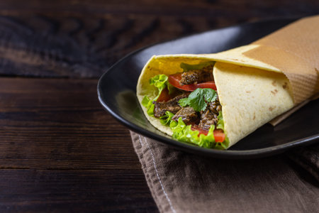 Burrito with slices of fried beef, tomato, lettuce, wrapped in cheese tortilla on black plate....