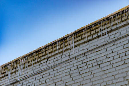 Roof of old building covered with sharp icicles. Large icicles hanging from the roof of an old brick building. Ice stalactite. Poor roof insulation leads to icicle formationの写真素材