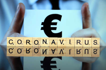 WROCLAW, POLAND - MARCH 30, 2020: The word CORONAVIRUS made of wooden letters, and man holding a business card with euro symbol in the background. Coronavirus (COVID-19) global disease 2020.のeditorial素材