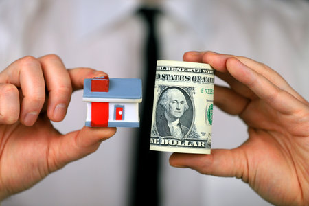A businessman in a white shirt and black tie holds a bundle of dollars and a miniature house in his hands.の写真素材