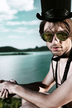 Fashion shot of a handsome young man posing topless in top hat and bow tie over beautiful landscape.の写真素材