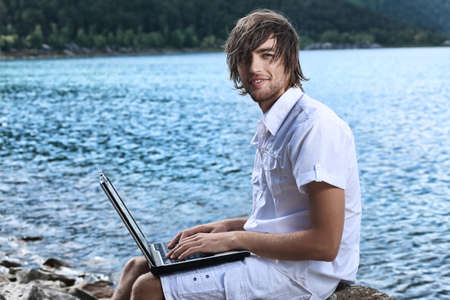 Happy young man sitting near the sea with a laptop.の写真素材