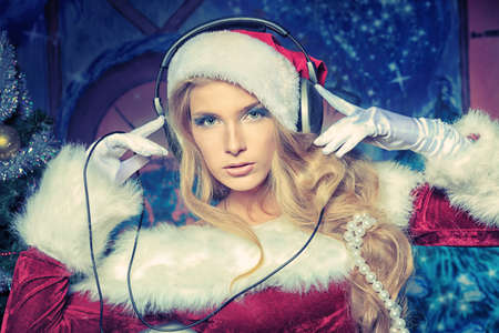 Beautiful young woman in Santa Claus clothes and headphones over Christmas background.の写真素材