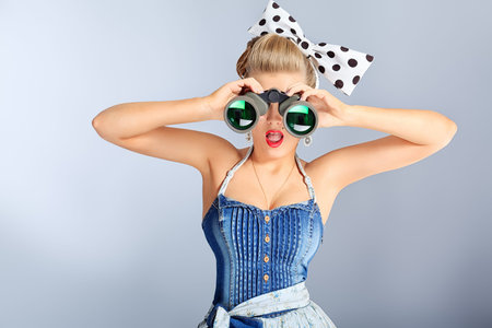 Beautiful young woman with pin-up make-up and hairstyle looking through binoculars.
