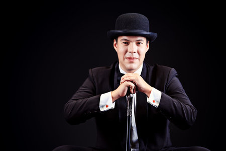 Portrait of an elegant man in black suit and black pot hat holding walking stick. Black background.