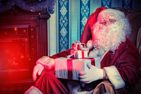 Santa Claus with gifts sitting in a comfortable chair near the fireplace at home. の写真素材