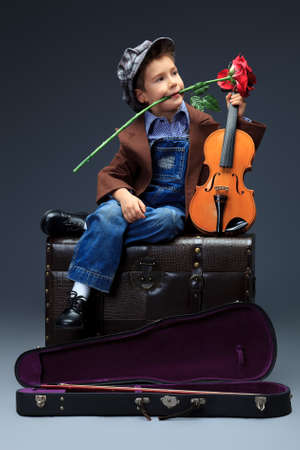 Portrait of a little boy sitting on a big old trunk with his violin and red rose. Retro style.の写真素材