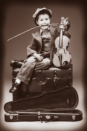 Portrait of a little boy sitting on a big old trunk with his violin and red rose. Retro style.