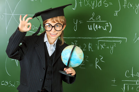 Portrait of a schoolboy in big round spectacles and academic hat at a classroom 
