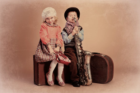 Cute little boy with his old saxophone is sitting on a suitcase with charming little lady. Retro...