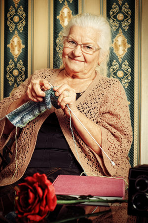 Portrait of a smiling senior woman knitting on spokes at home. Old-fashioned style.