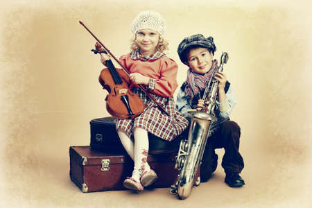 Cute little boy with his old saxophone is sitting on a suitcase with charming little lady violinist. Retro style.の写真素材