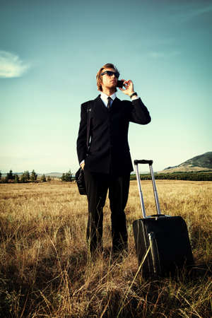 Handsome business man standing in a field with his suitcase and cell phone.の写真素材
