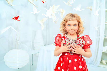 Cute little girl standing in a white room surrounded with paper birds and holding toy heart. の写真素材