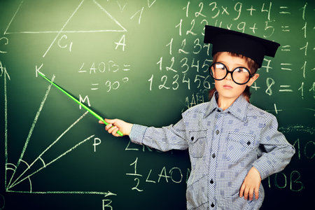 Portrait of a boy in round glasses and academic hat standing near the blackboard in a classroom.の写真素材