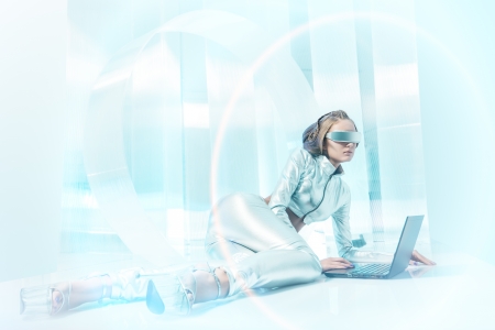 Beautiful young woman in silver latex costume with futuristic hairstyle and make-up working on a laptop. Sci-fi style.の写真素材