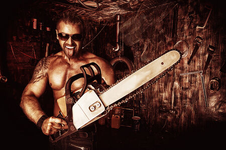 Expressive handsome muscular man with a chainsaw in the old garage.の写真素材