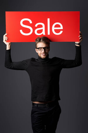 Modern handsome young man posing with a sign that says "SALE", over dark background.の写真素材