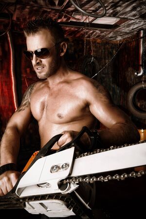 Expressive handsome muscular man with a chainsaw in the old garage.の写真素材