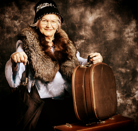 Beautiful old lady in an elegant old-fashioned clothes sitting on suitcases. Vintage style.の写真素材