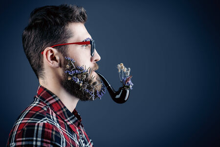 Handsome young man in glasses and a beard of flowers smoking a pipe. Profile portrait.の写真素材
