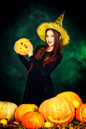 Pretty teen girl in a costume of witch standing with pumpkins over dark smoky background. Halloween.の写真素材