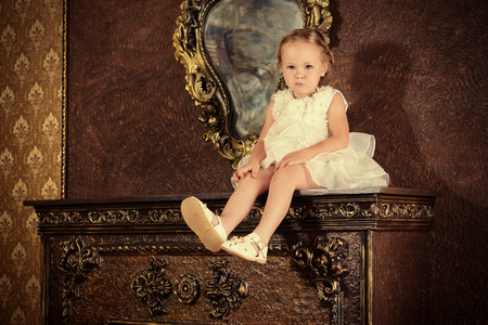 Cute little girl in a beautiful white dress sitting on the fireplace in the room. Vintage style.の写真素材