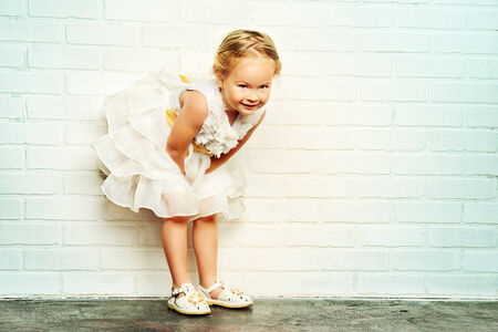 Angelic little girl in a beautiful white dress standing by the white brick wall. Childhood.の写真素材