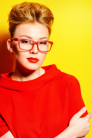 Close-up portrait of a stunning female model in red dress and elegant spectacles posing over yellow background. Beauty, fashion, optics.の写真素材