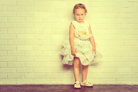 Angelic little girl in a beautiful white dress standing by the white brick wall. Childhood.の写真素材