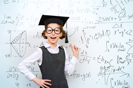 Happy smart schoolgirl in big glasses and academic hat performs the task at the blackboard....